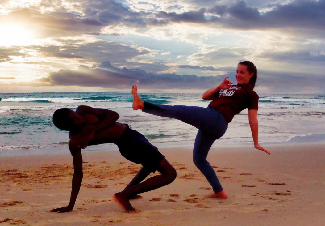 Zwei Personen am Strand, die Frau macht eine große kreisende Bewegung mit einem Bein, der Mann beugt sich zum Ausweichen nach hinten