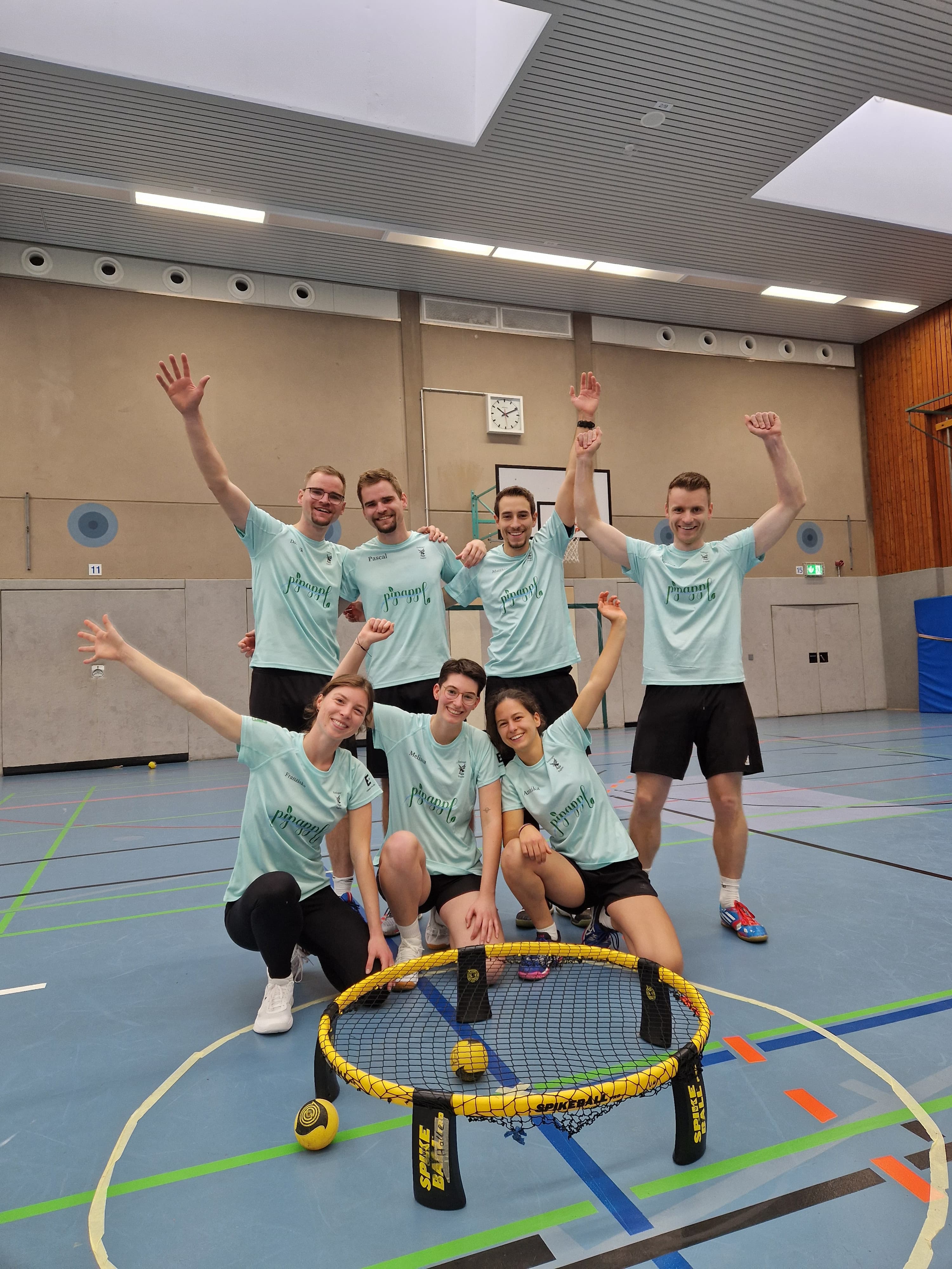 Sieben jubelnde Spielerinnen und Spieler posieren hinter einem Roundnet mit Ball